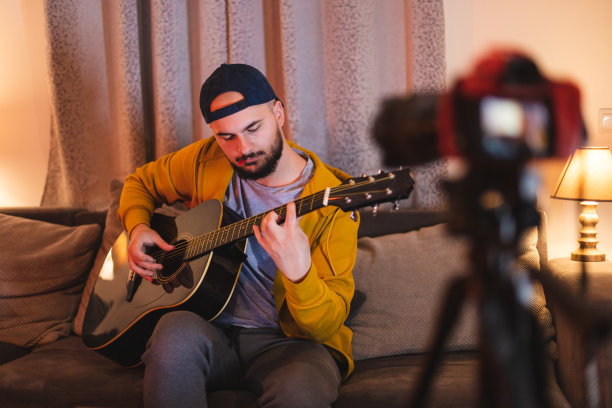 一个留着大胡子的年轻人一边在家里弹吉他，一边用单反相机自拍图片下载