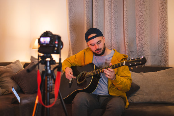 一个留着大胡子的年轻人一边在家里弹吉他，一边用单反相机为YouTube录制教程图片下载
