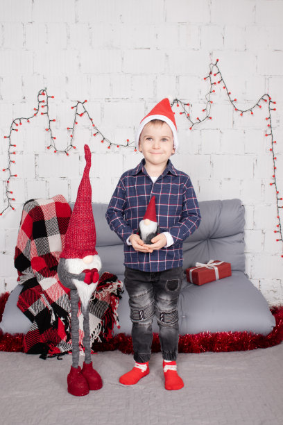 学龄前的小男孩戴着圣诞帽，站在高高的圣诞侏儒玩具旁边，手里拿着小矮人玩具。在灰色的房间里。圣诞快乐，新年快乐。圣诞装饰和玩具图片下载