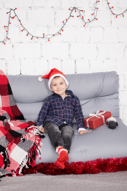 学龄前的小男孩戴着圣诞帽，站在高高的圣诞侏儒玩具旁边，手里拿着小矮人玩具。在灰色的房间里。圣诞快乐，新年快乐。圣诞装饰和玩具图片下载