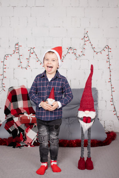 学龄前的小男孩戴着圣诞帽，站在高高的圣诞侏儒玩具旁边，手里拿着小矮人玩具。在灰色的房间里。圣诞快乐，新年快乐。圣诞装饰和玩具图片下载