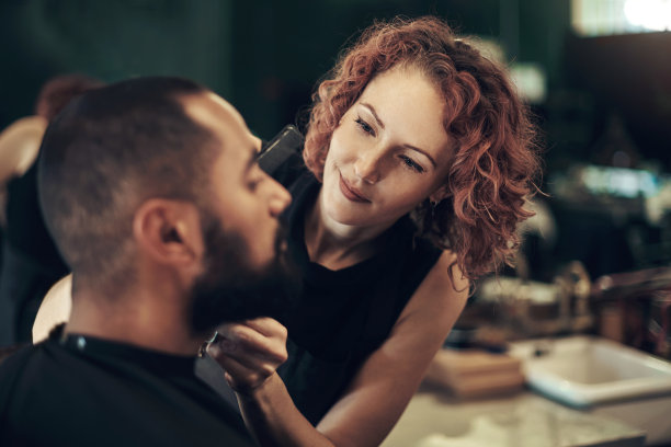 这张照片是一个漂亮的年轻美发师站在她的沙龙里给她的客户做胡须造型图片下载