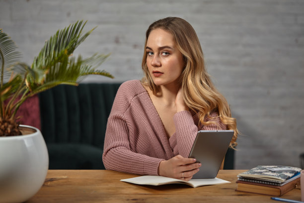 穿着粉色开襟羊毛衫的女博主拿着平板电脑。坐在木桌旁，桌上放着笔记本和一棵棕榈树。学生,博主。特写镜头图片下载