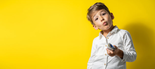 前视图是一个四岁的白人小男孩，穿着白色的衬衫，站在黄色的背景前面，拿着遥控器摆姿势，自信地看着镜头图片下载