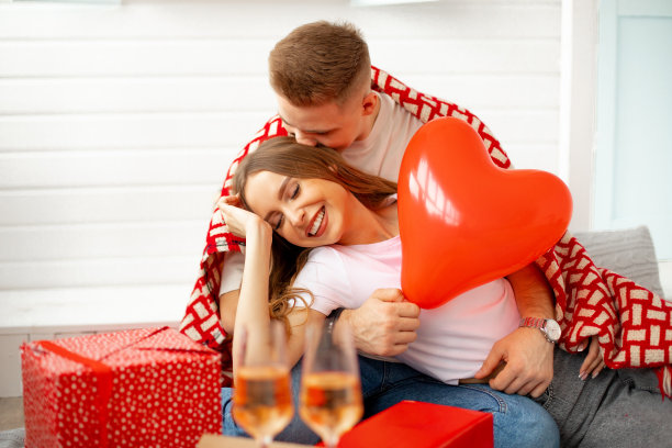 年轻人拥抱并亲吻他的爱人。他们在浪漫的家中用空灵的红心、礼物和红酒来庆祝情人节。爱和关心。美妙的时刻。图片下载