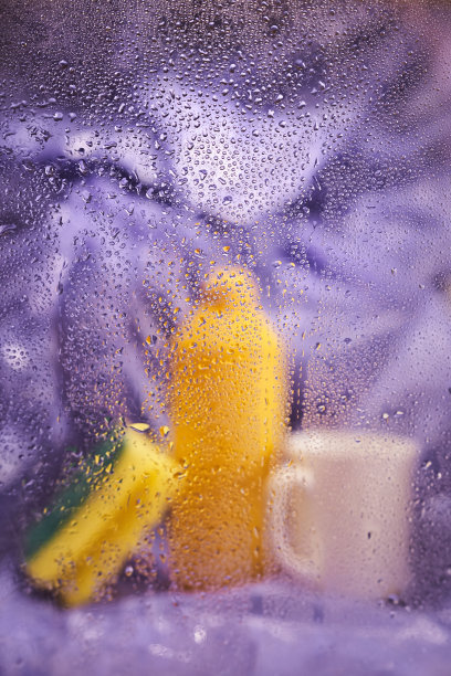 家用清洁产品和厨房配件。一个塑料杯和一个淡紫色背景上有海绵的瓶子。透过湿玻璃观看图片下载