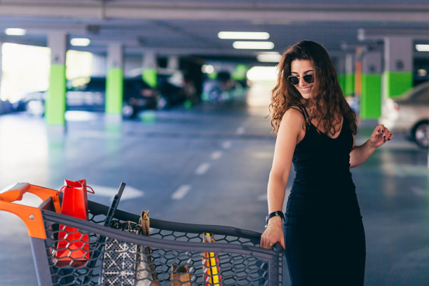 年轻漂亮的女子推着购物车，拿着装着商品和物品的包裹，来到购物中心地下停车场的汽车前图片下载