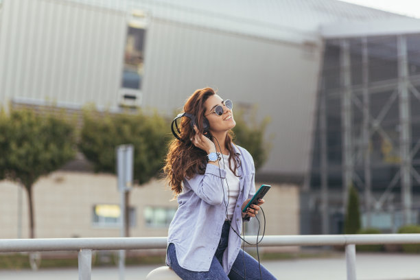 一个年轻美丽的女人的肖像，快乐地听音乐从手机在耳机里，坐在街上，附近的机场，火车站，公交车站图片下载