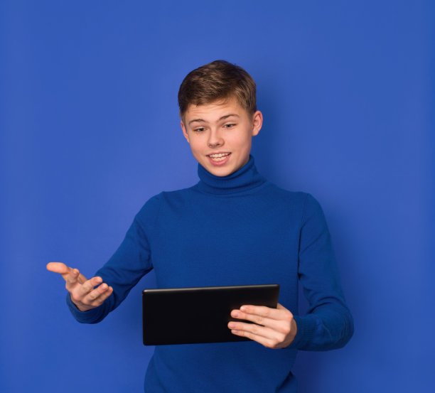 十几岁的男孩在蓝色背景下使用平板电脑。青少年、通讯、教育、科技的概念。图片下载