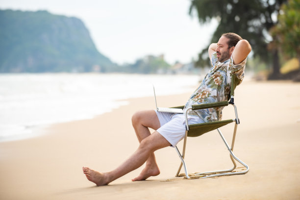 白人商人坐在户外的椅子上，在海滩上用笔记本电脑工作。图片下载