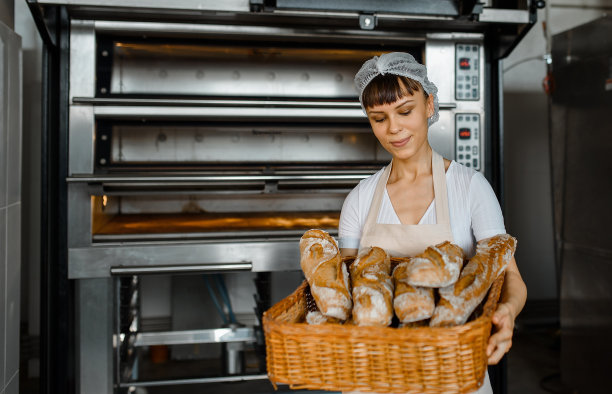 年轻的白人女面包师在烘焙工厂拿着一个装满新鲜面包的大篮子。图片下载