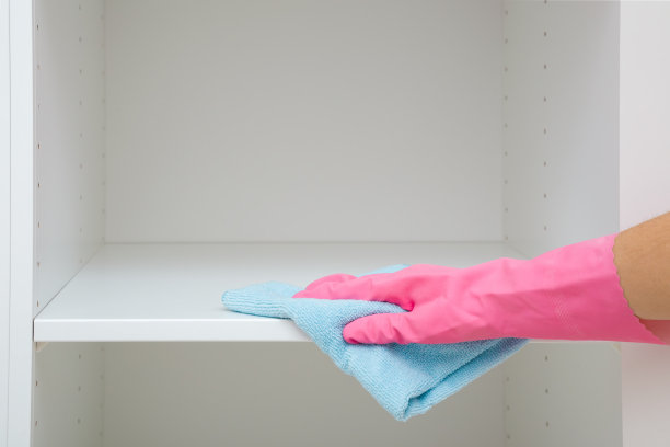 年轻的成年妇女戴着粉红色的橡胶防护手套，用蓝色的干抹布擦拭房间内木制衣柜内的白色搁板。特写镜头。前视图。在家定期打扫卫生。图片下载