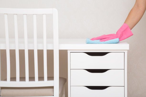 年轻女子手戴粉色橡胶保护手套，手里拿着蓝色干抹布，擦拭家里办公室有抽屉的白色工作台上的灰尘。特写镜头。房间的一般或定期清洁。清洁服务。图片下载