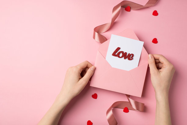 第一人称俯视图照片的情人节装饰女孩的手，与卡片和题字的粉红色信封，爱心缎子卷曲丝带在孤立的粉彩粉红色背景与copyspace图片下载
