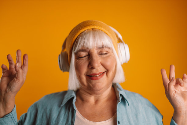 幸福的50多岁的女人戴着耳机听音乐唱歌曲录制声音，用手机录音机录在黄色背景上画室肖像。图片下载