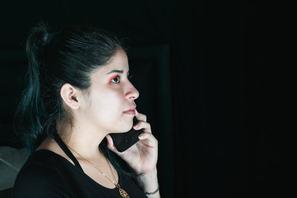 美丽的拉丁女人与绿色的头发，接受电话在她的手机，一种专注的表情。女孩坐在房间里和朋友聊天。非常暗的黑色背景。图片下载