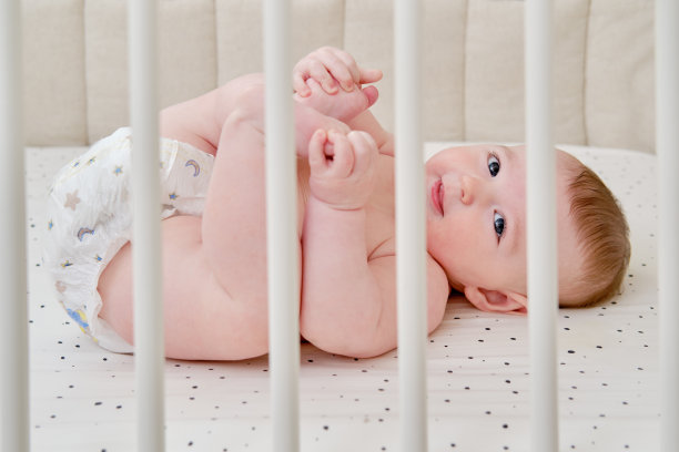 一个穿着尿布、长着牙齿的快乐婴儿把他的手放进嘴里。婴儿床里的滑稽孩子图片下载