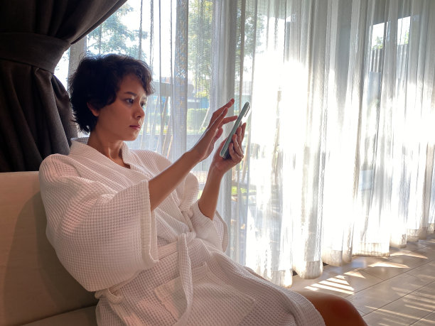 女人在家里或公寓里。穿着白色浴袍的女孩拿着智能手机和一杯咖啡站在大窗户前。，社交距离和在家工作的概念。图片下载