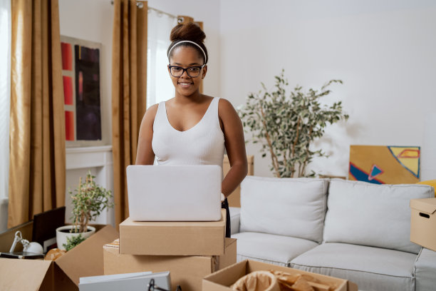 一个戴眼镜的女人微笑着站在一所公寓的中间，一个女孩在她的纸箱周围浏览着网上商店，为房子订购家具图片下载