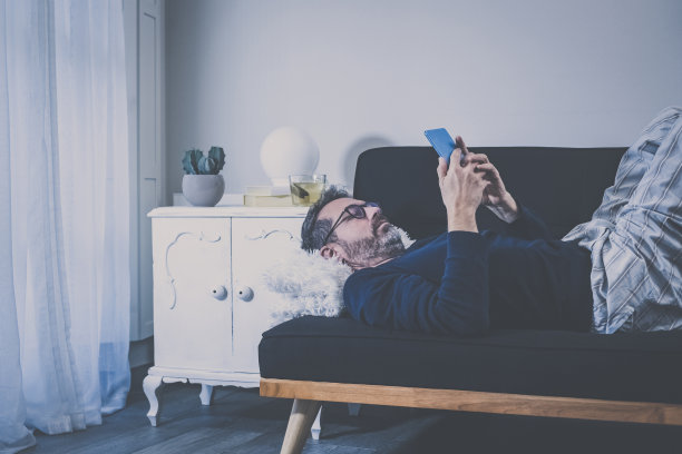 男人拿着手机和垫子躺在沙发上。沉迷于科技的男性享受自由时间。男孩在客厅里用在线设备放松。悠闲、休闲、生活方式的新传播理念图片下载