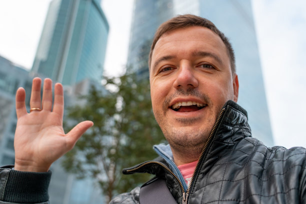 一个留着胡子的快乐的年轻人一边笑着一边自拍，一边在城市里享受一天的时光。年轻英俊的白人男子微笑着愉快地在他的旅行期间在城市步行相机自拍图片下载