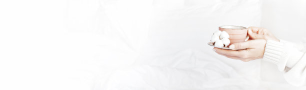 女人的手在白色的针织毛衣，杯中热茶茶托和精致的棉花花在床上。在家享受舒适的早餐。生活方式温和的背景Copy Space秋冬概念图片下载