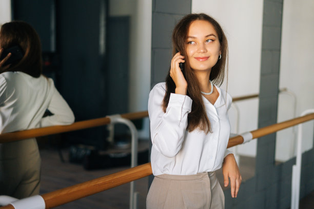微笑的年轻女子的肖像在时尚的衣服上讲手机站在房间与镜子，看向别处。图片下载