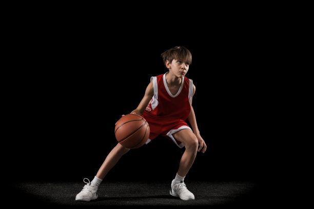 全长度水平工作室拍摄的少年在红色的制服打篮球，在黑色的背景下做运球练习图片下载