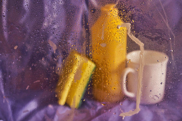 家用清洁产品和厨房配件。一个塑料杯和一个淡紫色背景上有海绵的瓶子。透过湿玻璃观看图片下载