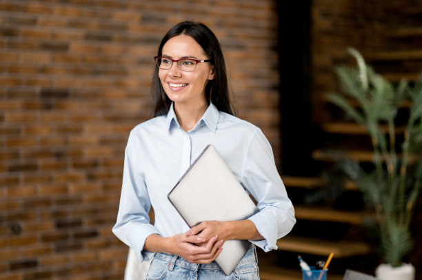 肖像美丽聪明自信的年轻成年黑发白人妇女，办公室经理，教师，经纪人戴眼镜，站在办公室，拿着笔记本电脑在她的手，看着一边，微笑图片下载