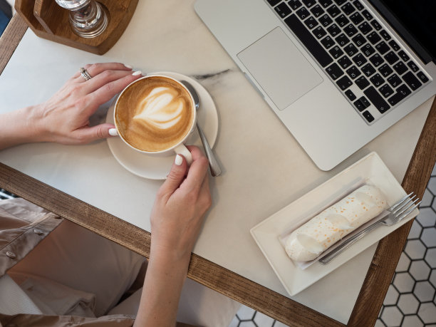 工作生活的平衡。在清晨的阳光下，一个女人在咖啡馆里用笔记本电脑工作，一边喝着咖啡，一边吃着泡芙。图片下载