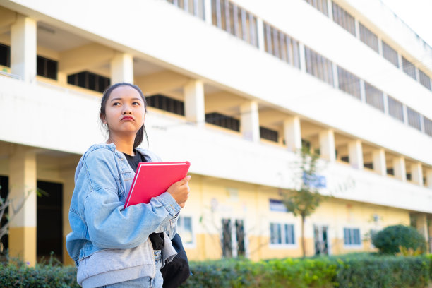 微笑的女孩拿着粉红色的书站在建筑的背景上，回到学校。图片下载
