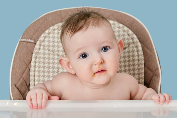 一个被食物弄脏的婴儿坐在给孩子喂食的高脚椅子上，画室的蓝色背景。孩子男孩在六个月大的时候坐在儿童椅上吃东西，复印空间图片下载