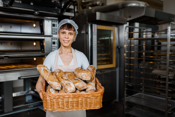 年轻的白人女面包师在烘焙工厂拿着一个装满新鲜面包的大篮子。图片下载