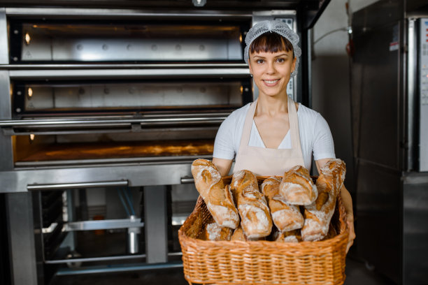 年轻的白人女面包师在烘焙工厂拿着一个装满新鲜面包的大篮子。图片下载