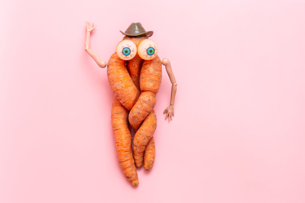 粉色背景上滑稽丑陋的扭曲胡萝卜。胡萝卜有手，眼睛和帽子。蔬菜的玩具图片下载