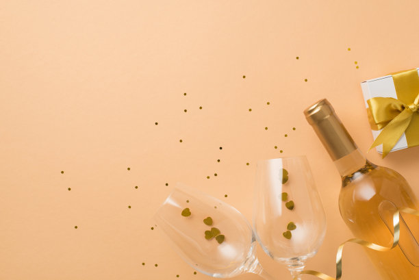 俯视图照片的情人节装饰白色礼盒与金色蝴蝶结酒瓶，两个葡萄酒杯与心形纸屑和亮片在孤立的粉彩米色背景与空空间图片下载