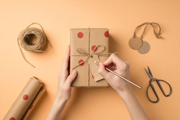 第一人称俯视图照片的手工工具，女孩的手写在标签附在牛皮纸礼品盒与圆点图案和缠绕蝴蝶结孤立的米色背景copyspace图片下载