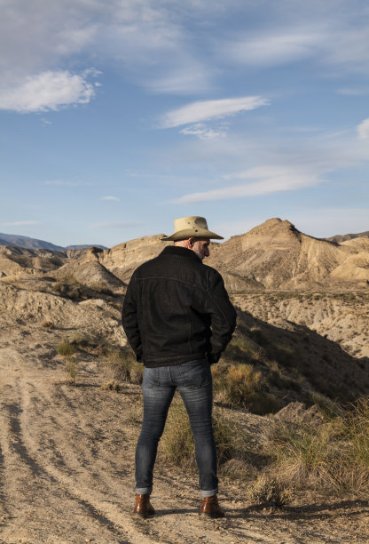戴着牛仔帽的成年男子站在塔伯纳斯沙漠的群山前。西班牙阿尔梅里亚图片下载