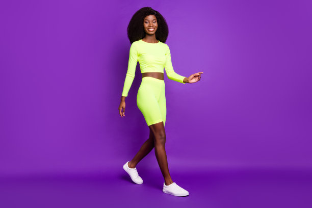 迷人的年轻女士走的照片看相机穿绿色衬衫短裤运动鞋孤立的紫色背景图片下载