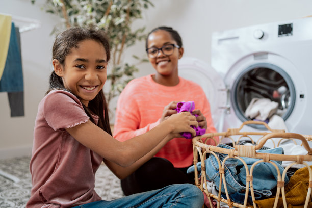 一名微笑的妇女和她的女儿从洗衣篮里拿出衣服，把它们装进洗衣机的桶里，女孩帮助做家务图片下载