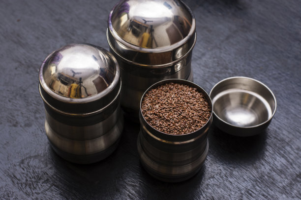 三个不锈钢容器，其中两个是封闭的，一个是打开的，装满了种子，放在黑色的木桌上。图片下载