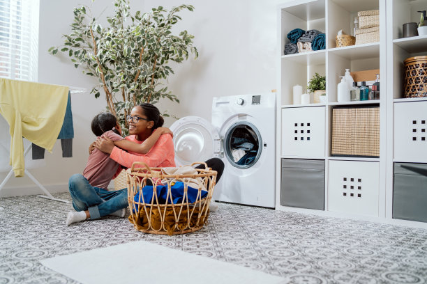 妈妈紧紧抱着女儿，坐在浴室地板上的洗衣间里，柳条篮子里装满了待洗的衣服，互相帮助做家务，亲情图片下载