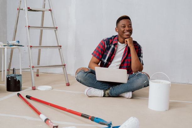 满意的黑皮肤男孩支持自己的下巴，坐在公寓的纸箱上，膝盖上，拿着笔记本电脑，寻找灵感，他周围正在翻新，躺着滚轴图片下载