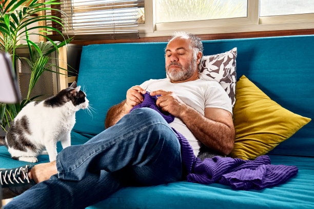 一名留着胡子的中年男子坐在沙发上，用毛线球编织针编织一条紫罗兰色的羊毛围巾，一边和他的猫玩耍，一边编织，这是他保持积极心理健康的爱好之一图片下载