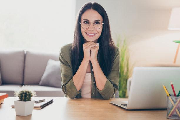 照片上漂亮积极的女孩坐在桌面后，双臂抱在下巴下，看着相机笑容满面地回家图片下载