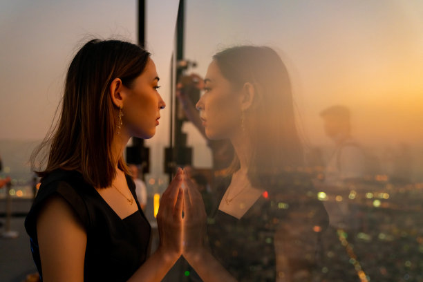 亚洲妇女的肖像站在窗口的摩天大楼和看着夜晚的城市景观。图片下载