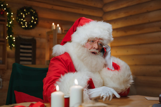 圣诞老人在木屋里舒适的装饰过的书房里打电话，在照片的前景中，我们看到他的桌子上放着蜡烛图片下载