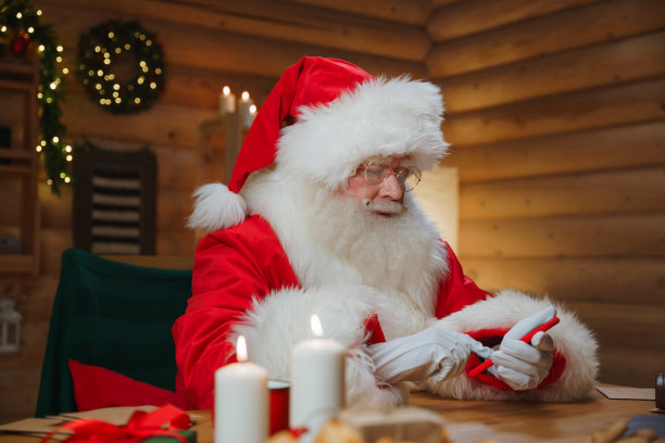 圣诞老人坐在摆满装饰品和蜡烛的书房里，用他的红色智能手机打字图片下载