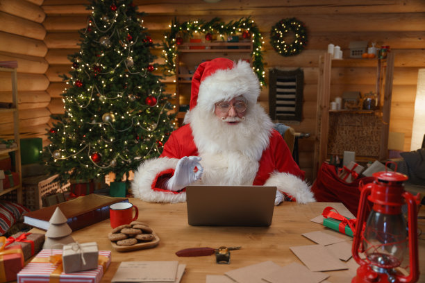 圣诞老人正坐在办公桌前，用笔记本电脑视频聊天。他的办公室装饰得很有圣诞气氛，身后摆着一棵漂亮的圣诞树图片下载